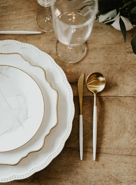 A close-up of white and gold charger plates, dinnerware, and flatware arranged on a wooden table.
