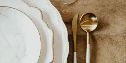 A close-up of white and gold charger plates, dinnerware, and flatware arranged on a wooden table.