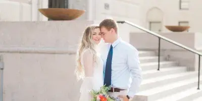 A professional photograph of a bride and groom standing together outdoors in front of a stone building.
