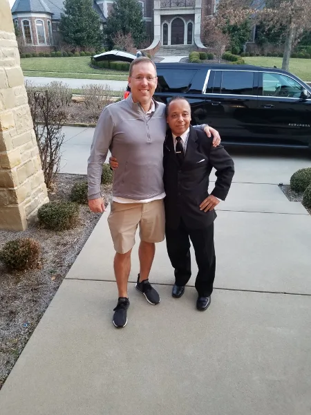 A professional chauffeur in a suit posing with a client in front of a black SUV.