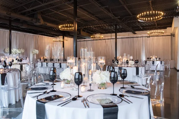 A spacious banquet hall with round tables set for a formal event, featuring white tablecloths, black napkins, and tall floral centerpieces under modern chandeliers.