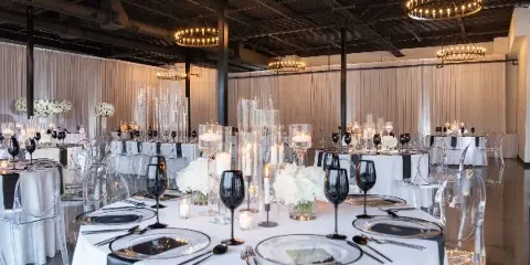 A spacious banquet hall with round tables set for a formal event, featuring white tablecloths, black napkins, and tall floral centerpieces under modern chandeliers.