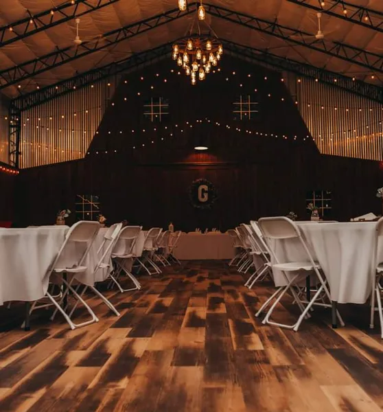 Interior of a rustic event barn featuring a wooden dance floor, string lights, and white reception tables.