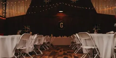 Interior of a rustic event barn featuring a wooden dance floor, string lights, and white reception tables.