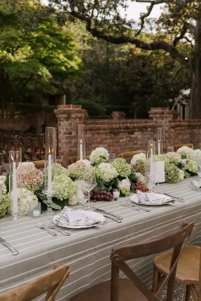 A long outdoor dining table set with a striped tablecloth, white and pink hydrangea floral centerpieces, tall glass candle holders, and silverware.
