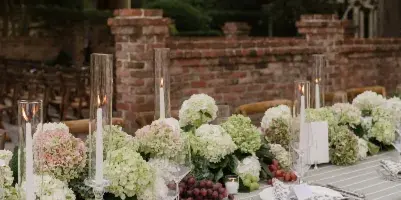A long outdoor dining table set with a striped tablecloth, white and pink hydrangea floral centerpieces, tall glass candle holders, and silverware.