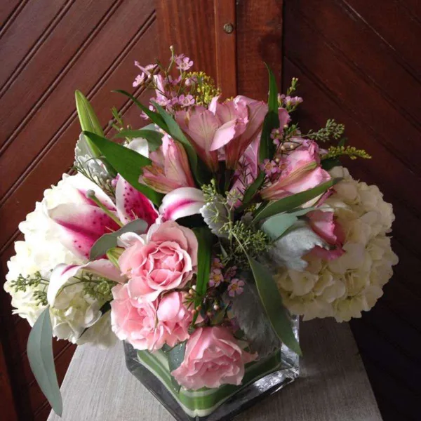 A floral arrangement with pink lilies, pink roses, and white hydrangeas in a square glass vase.