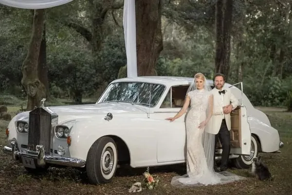A bride and groom standing next to a white vintage Rolls-Royce in a wooded outdoor setting.