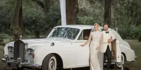 A bride and groom standing next to a white vintage Rolls-Royce in a wooded outdoor setting.