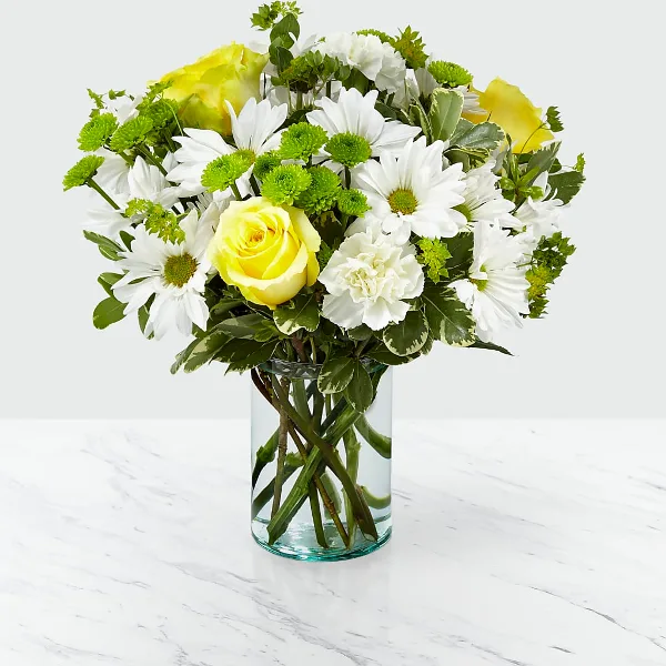 A fresh flower arrangement featuring white daisies, yellow roses, and green filler flowers in a clear glass vase.