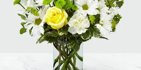 A fresh flower arrangement featuring white daisies, yellow roses, and green filler flowers in a clear glass vase.