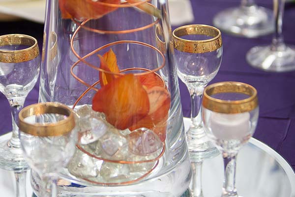 A close-up of an event table setting featuring a glass vase centerpiece with copper wire and orange flowers, surrounded by gold-rimmed glassware on a purple tablecloth.