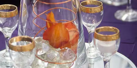 A close-up of an event table setting featuring a glass vase centerpiece with copper wire and orange flowers, surrounded by gold-rimmed glassware on a purple tablecloth.