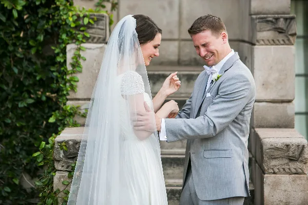A bride and groom share an emotional first-look moment outdoors, with the groom smiling and the bride looking down at their joined hands.