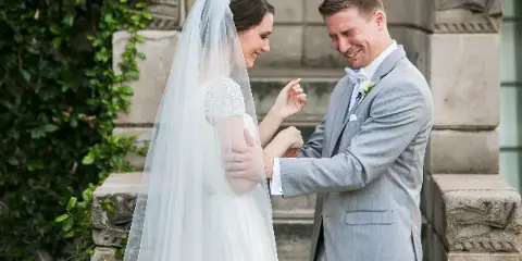 A bride and groom share an emotional first-look moment outdoors, with the groom smiling and the bride looking down at their joined hands.