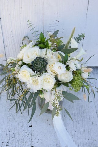 A white bridal bouquet featuring roses, succulents, and greenery, resting on a white wooden surface.