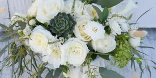 A white bridal bouquet featuring roses, succulents, and greenery, resting on a white wooden surface.
