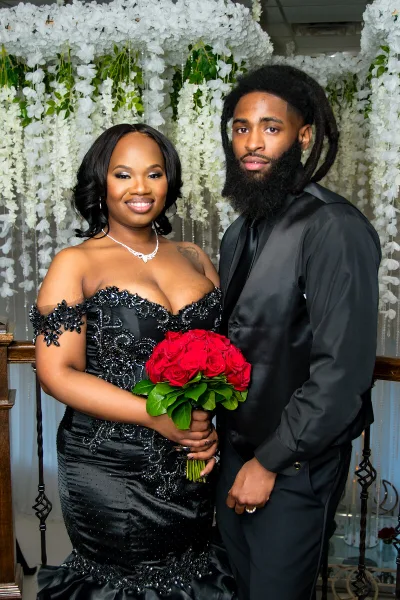 A professional wedding portrait of a bride in a black gown holding a red rose bouquet and a groom in a black suit, standing together in front of a white floral backdrop.