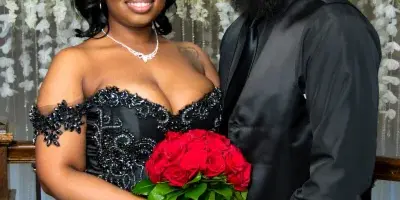 A professional wedding portrait of a bride in a black gown holding a red rose bouquet and a groom in a black suit, standing together in front of a white floral backdrop.
