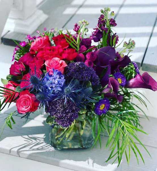 A vibrant floral arrangement in a glass vase featuring pink roses, purple calla lilies, and blue thistles.