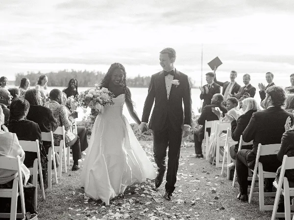 A black and white photograph of a bride and groom walking down the aisle during an outdoor wedding ceremony with seated guests.