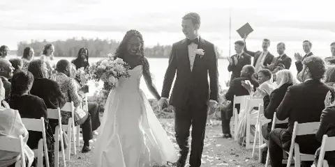 A black and white photograph of a bride and groom walking down the aisle during an outdoor wedding ceremony with seated guests.