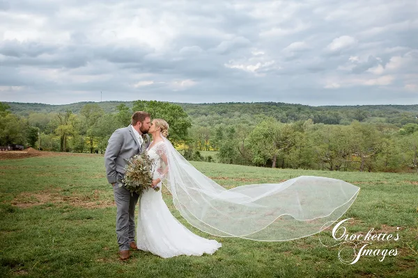 A bride and groom kissing in a grassy field with a long veil blowing in the wind, set against a backdrop of rolling hills and trees.