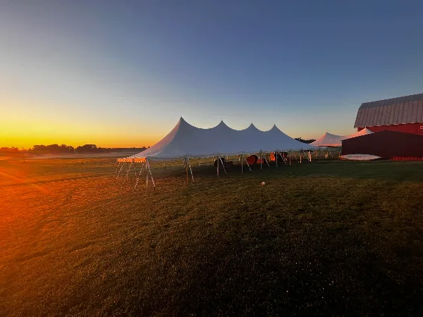 A large white event tent with multiple peaks set up in a grassy field at sunset.