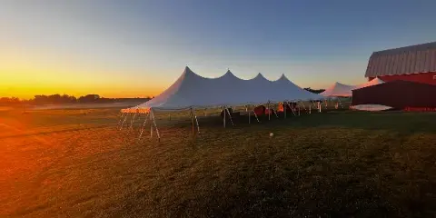 A large white event tent with multiple peaks set up in a grassy field at sunset.