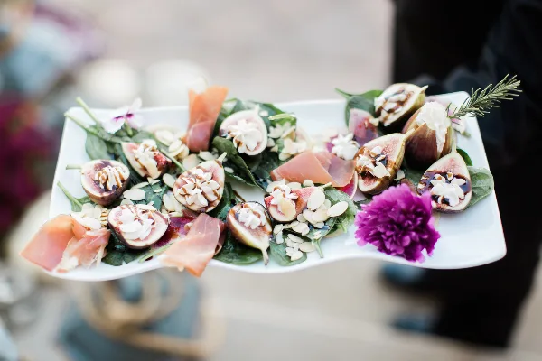 A white rectangular platter holding a gourmet salad with sliced figs, prosciutto, cheese, and a purple flower garnish.