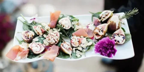 A white rectangular platter holding a gourmet salad with sliced figs, prosciutto, cheese, and a purple flower garnish.