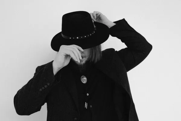Black and white studio portrait of a man with a beard wearing a black hat and coat, adjusting his hat.