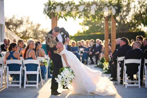 A groom dipping his bride for a kiss during an outdoor wedding ceremony under a floral arch.