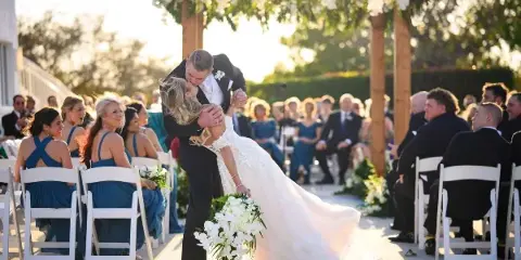 A groom dipping his bride for a kiss during an outdoor wedding ceremony under a floral arch.