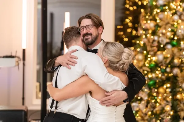A wedding DJ in a tuxedo hugging a bride and groom at a wedding reception.