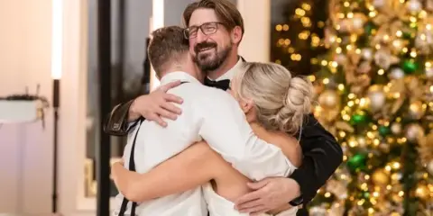 A wedding DJ in a tuxedo hugging a bride and groom at a wedding reception.