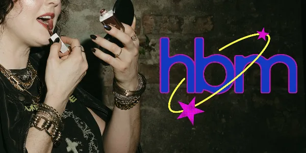 A person applying lip makeup while holding a compact mirror, with the 'hbm' logo overlaid.