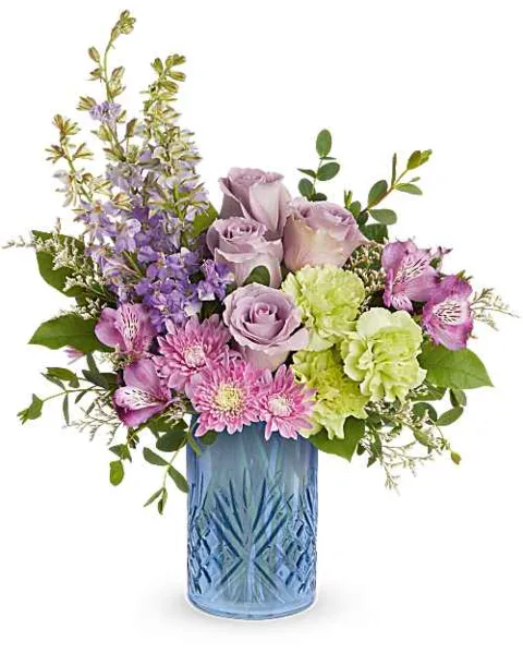 A mixed flower arrangement featuring purple roses, pink daisies, and green hydrangeas in a blue glass vase.