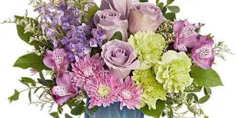 A mixed flower arrangement featuring purple roses, pink daisies, and green hydrangeas in a blue glass vase.