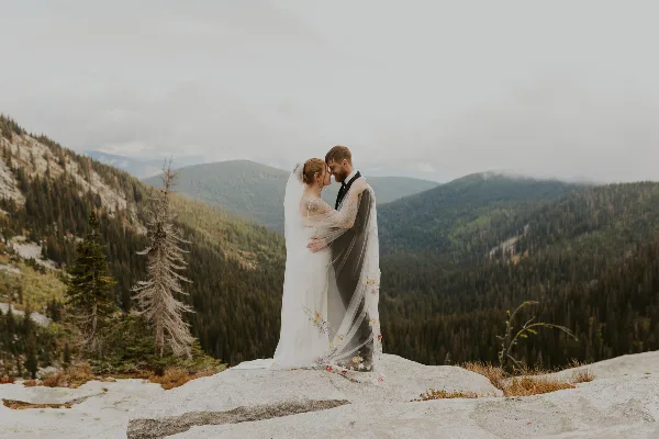A bride and groom embracing on a rocky mountain overlook with a forested valley in the background.