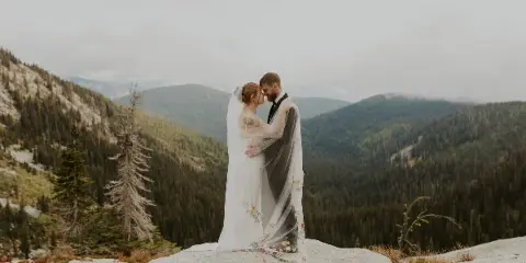 A bride and groom embracing on a rocky mountain overlook with a forested valley in the background.