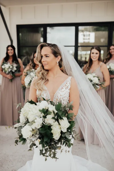 A bride with styled hair and makeup holding a bouquet, with bridesmaids in the background.
