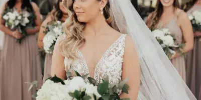 A bride with styled hair and makeup holding a bouquet, with bridesmaids in the background.