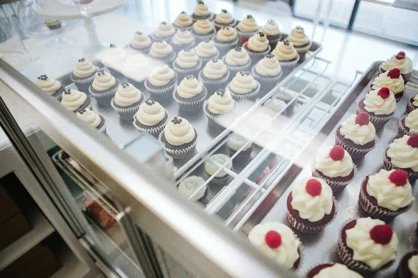 A bakery display case filled with rows of decorated cupcakes, including chocolate cupcakes with white frosting and red velvet cupcakes topped with raspberries.