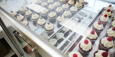 A bakery display case filled with rows of decorated cupcakes, including chocolate cupcakes with white frosting and red velvet cupcakes topped with raspberries.