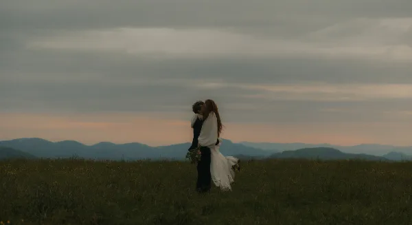 A bride and groom embracing in a field with mountains in the background at dusk.