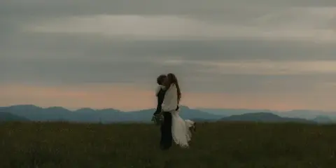 A bride and groom embracing in a field with mountains in the background at dusk.