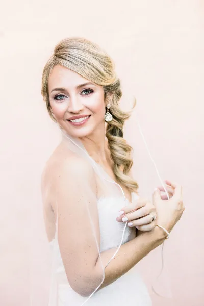 A smiling blonde bride in a white wedding dress and veil posing against a soft pink background.