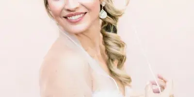 A smiling blonde bride in a white wedding dress and veil posing against a soft pink background.