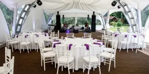 A large white event tent set up outdoors with round tables, white chairs, and a stage with audio equipment.